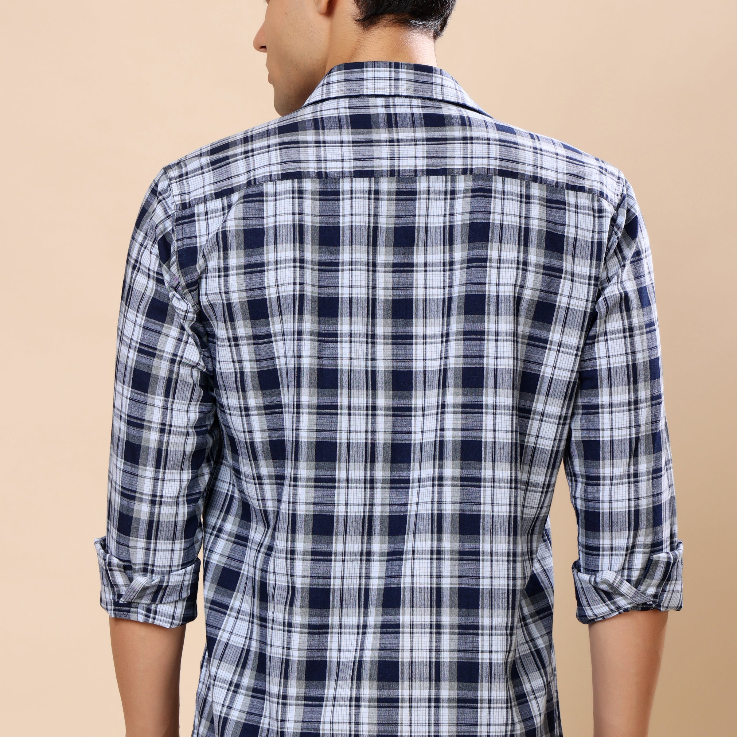 A man viewed from the back wearing a navy blue and white plaid long-sleeve shirt with rolled-up sleeves, set against a solid beige background.