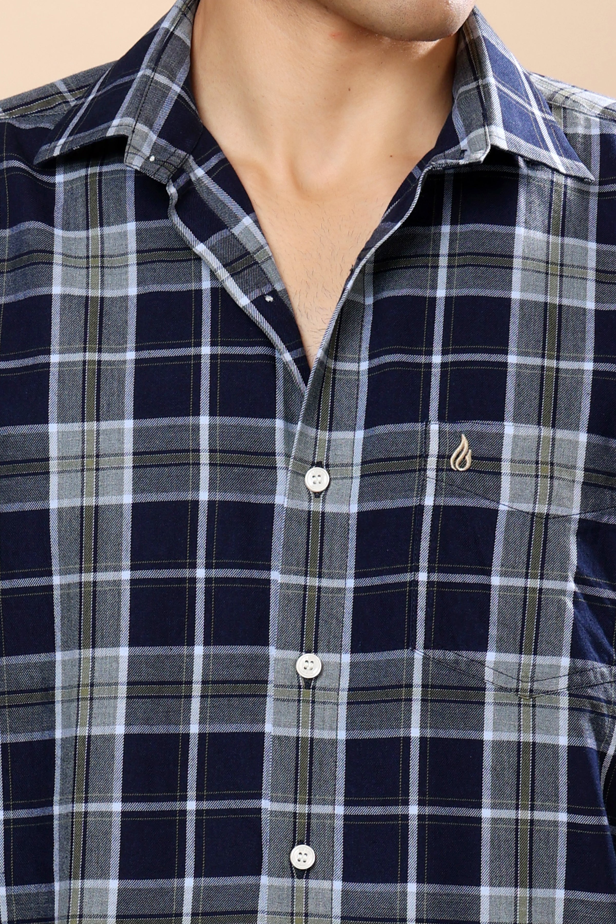 A close-up of a men's plaid button-down shirt worn by a model. The shirt features a navy blue, gray, and white checkered pattern with a small gold embroidered flame logo on the left chest.