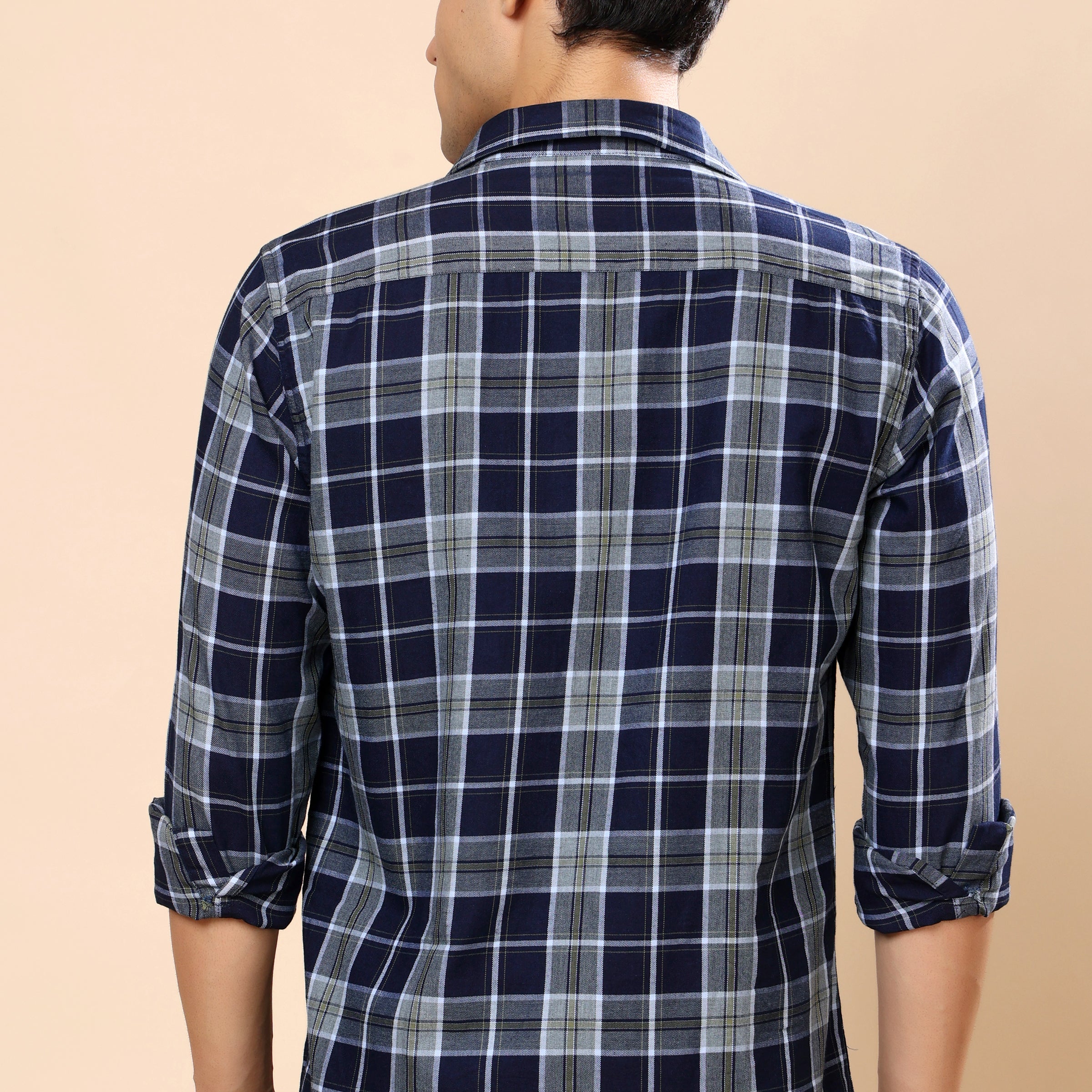A man viewed from the back wearing a navy blue and gray plaid long-sleeve shirt with rolled-up sleeves, set against a solid beige background.
