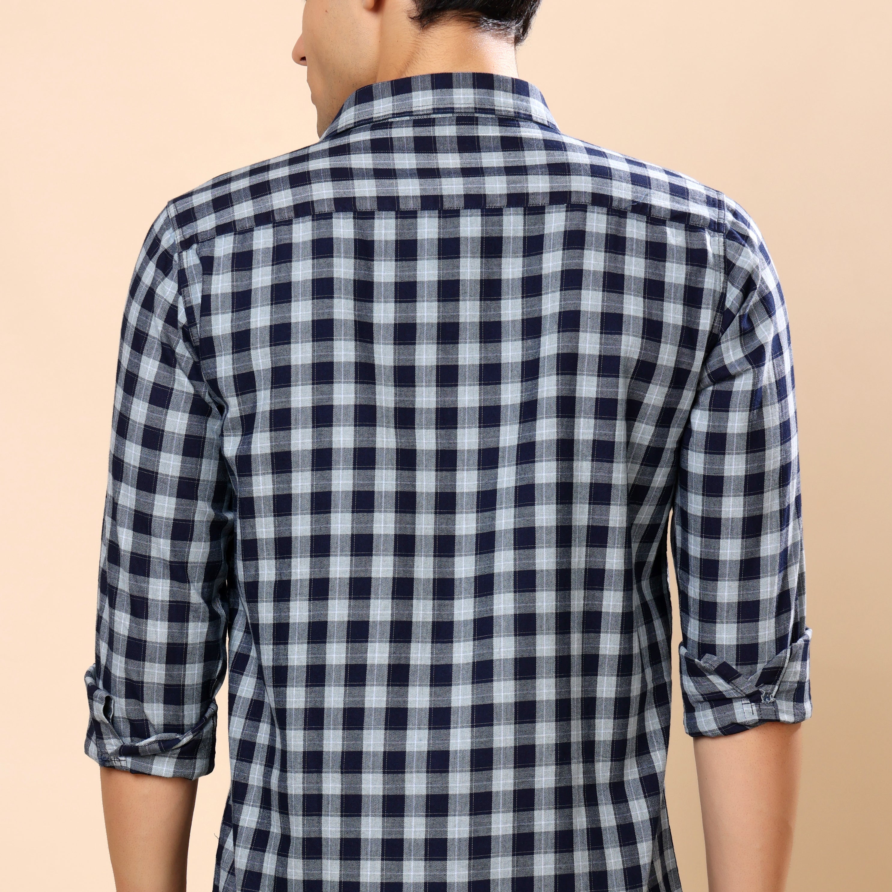 A man viewed from the back wearing a navy and white checkered long-sleeve shirt with rolled-up sleeves, paired with dark gray jeans, against a solid beige background.