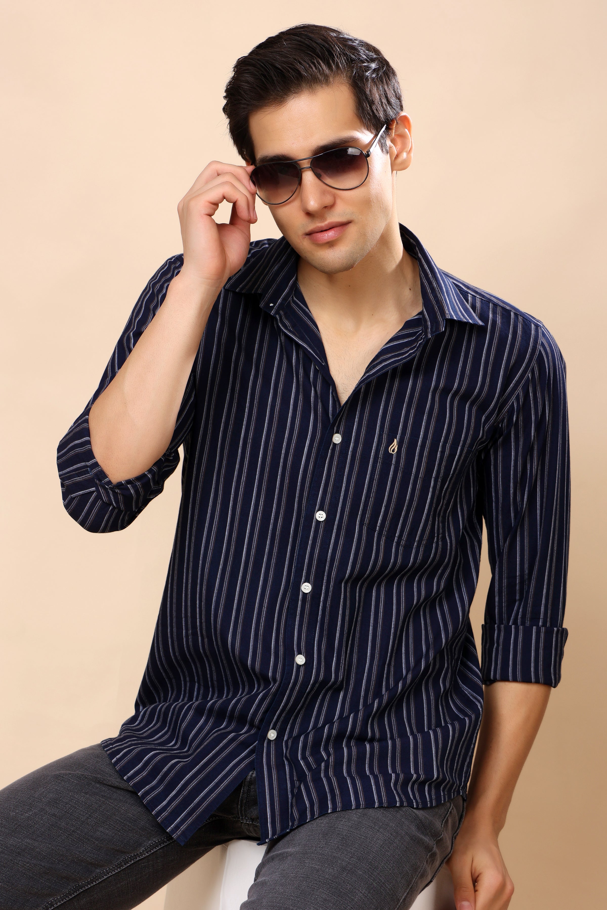 A man wearing a navy blue and white vertically striped button-down shirt with dark gray jeans, sitting against a solid beige background. He is wearing dark sunglasses and adjusting them with his right hand.