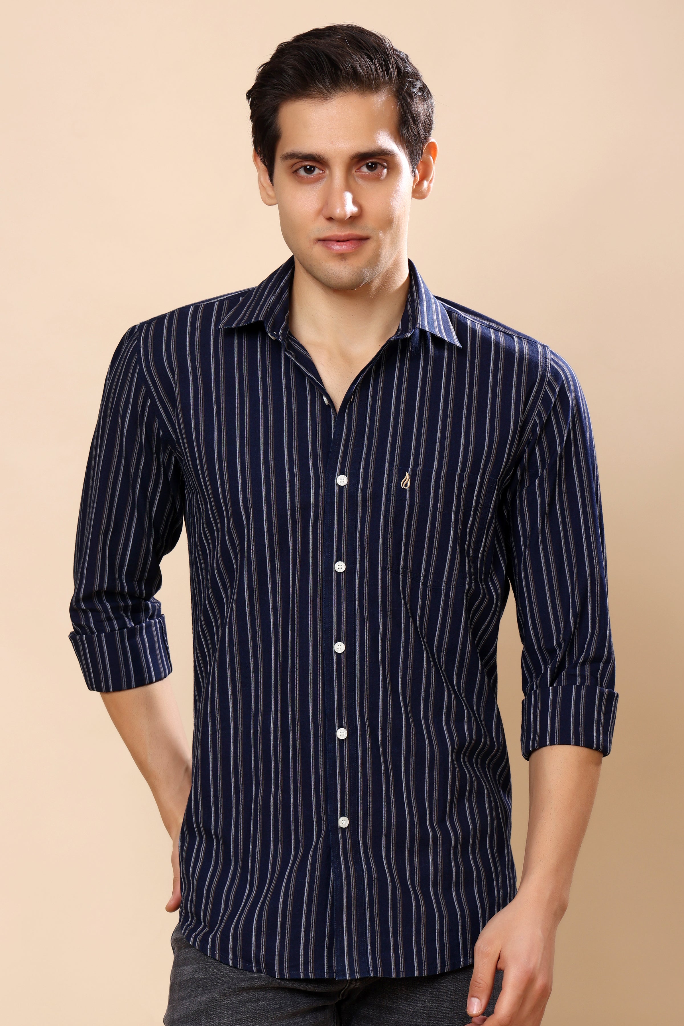 A man wearing a navy blue long-sleeve button-up shirt with thin white vertical stripes, standing against a solid beige background.