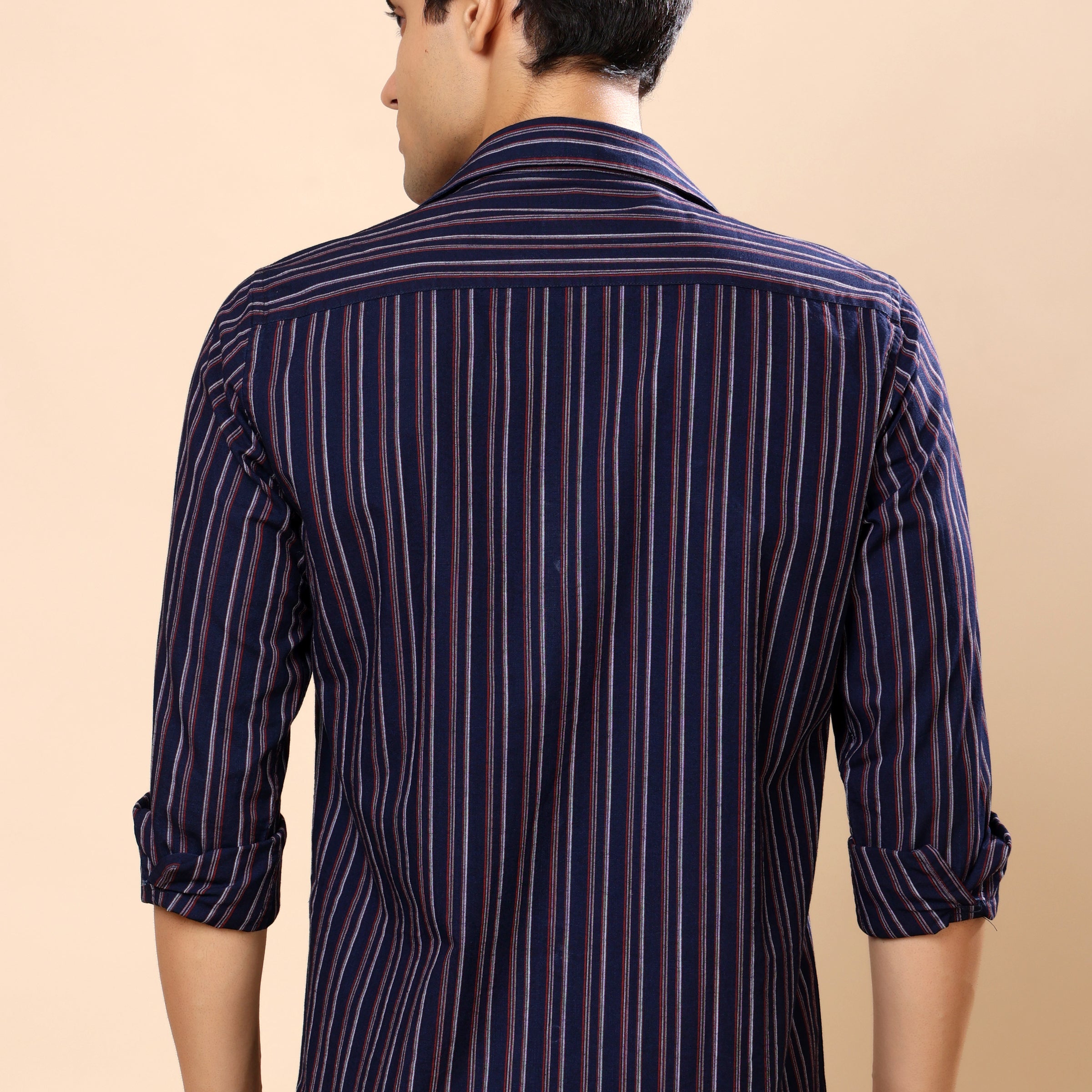 A man viewed from the back wearing a navy blue striped button-down shirt with thin red and white vertical lines, set against a solid beige background.