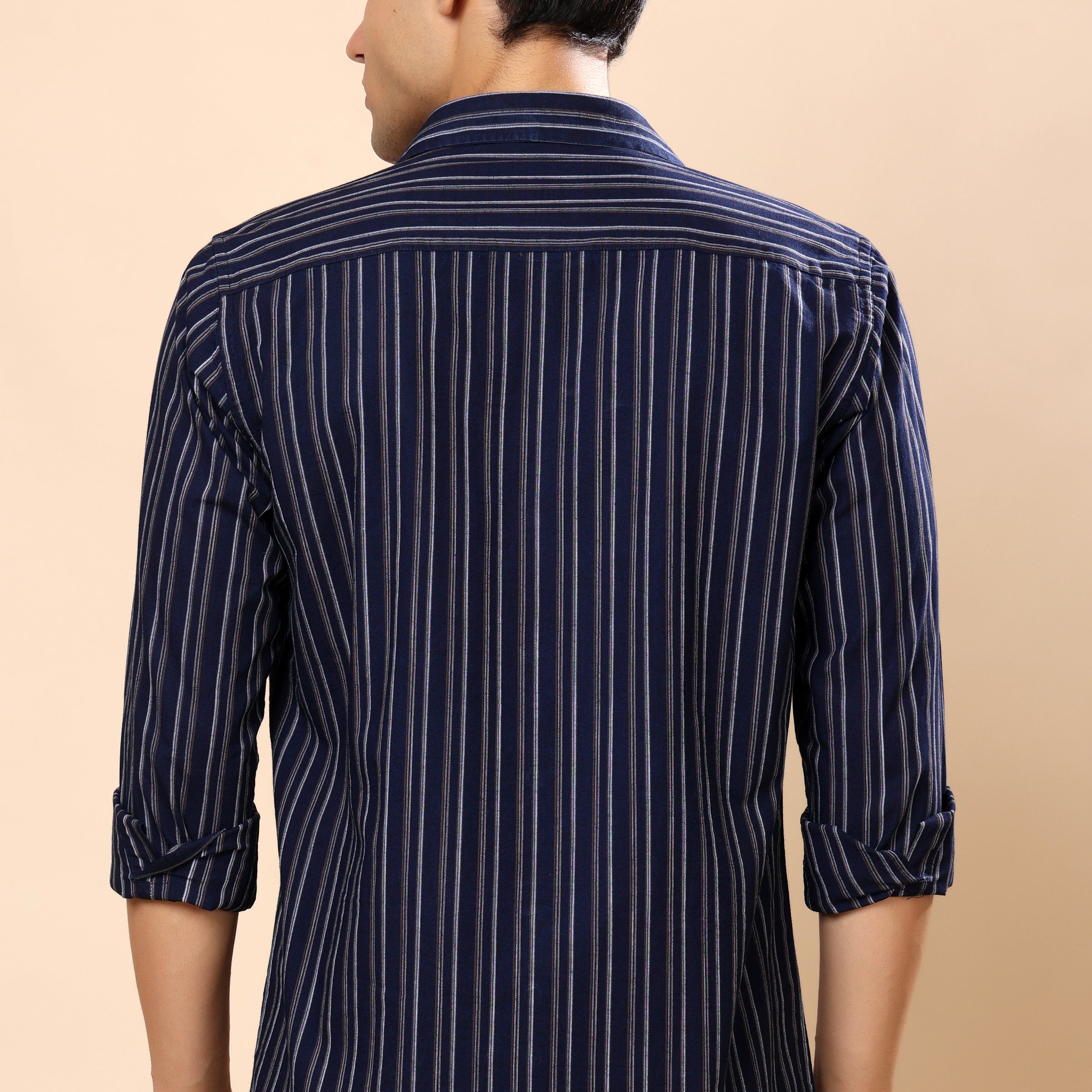 A man viewed from the back wearing a navy blue and white vertically striped long-sleeve shirt, paired with dark gray jeans, against a solid beige background.