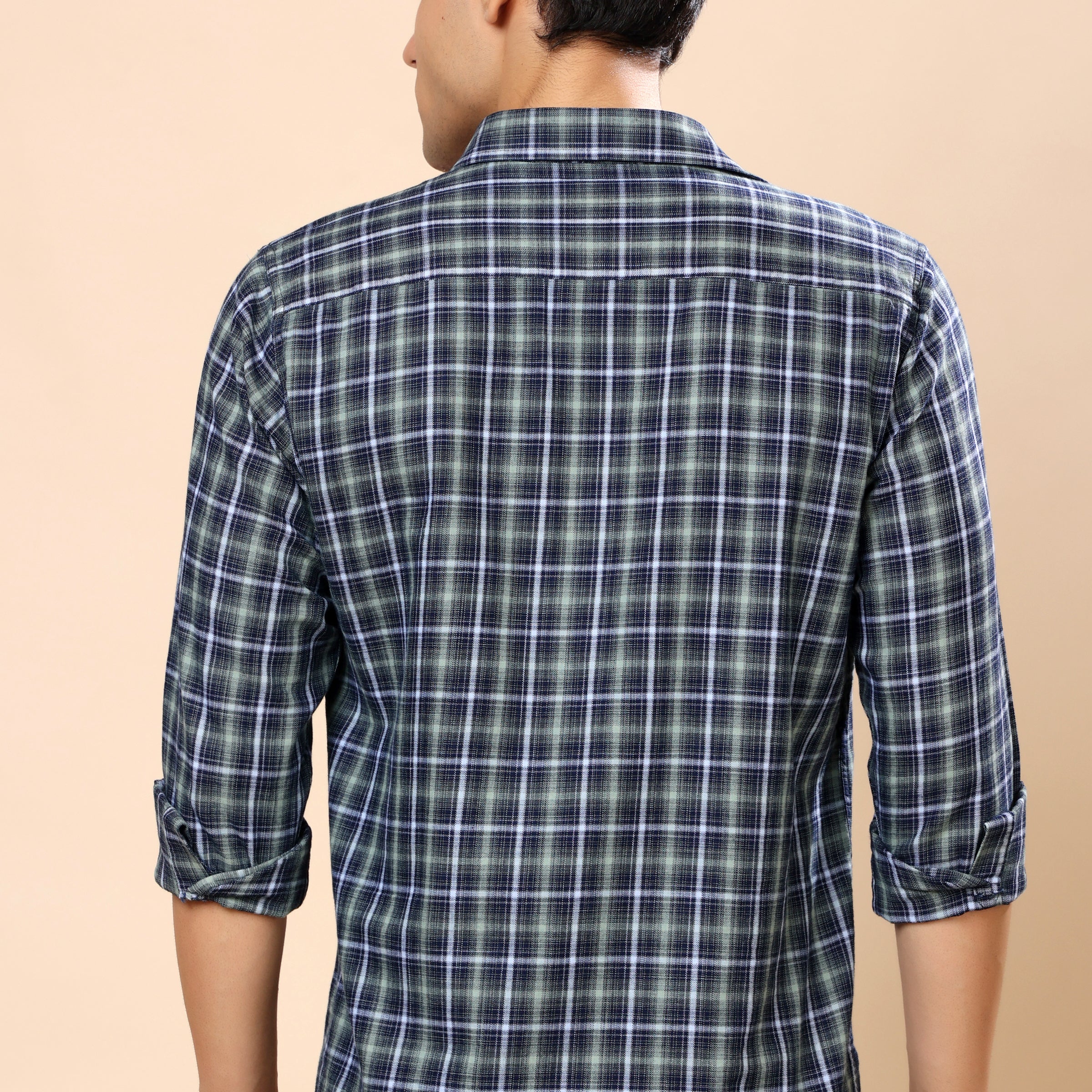 A man viewed from the back wearing a long-sleeve plaid button-up shirt in dark blue and white, paired with dark gray jeans, against a solid beige background.