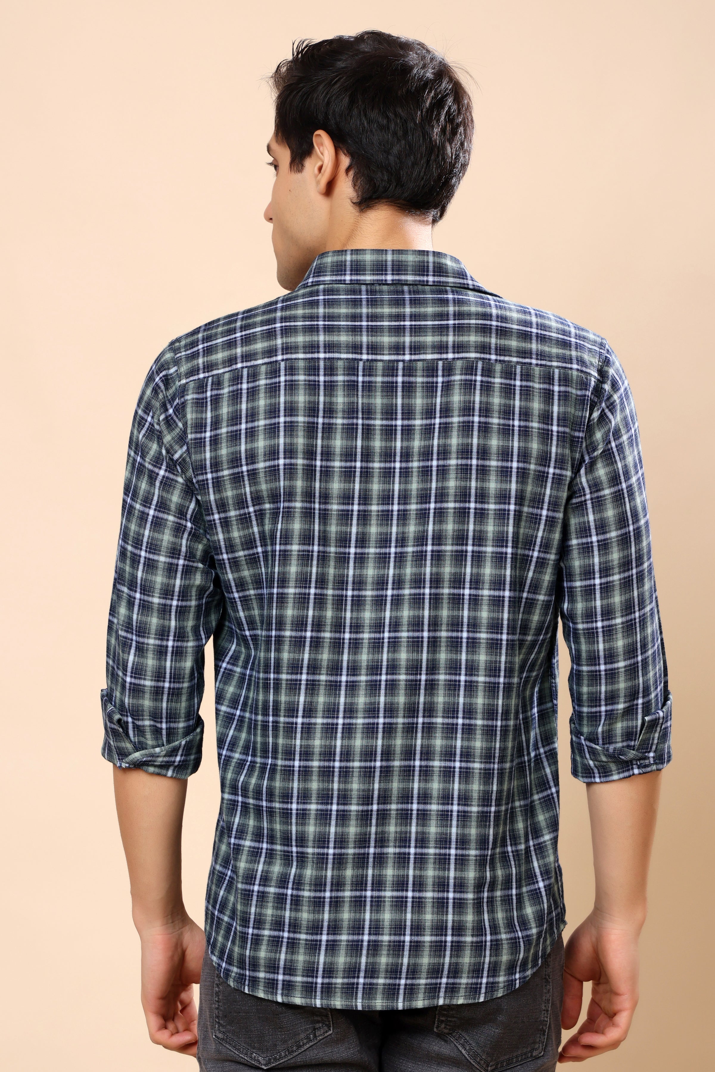 A man viewed from the back wearing a long-sleeve plaid button-up shirt in dark blue and white, paired with dark gray jeans, against a solid beige background.