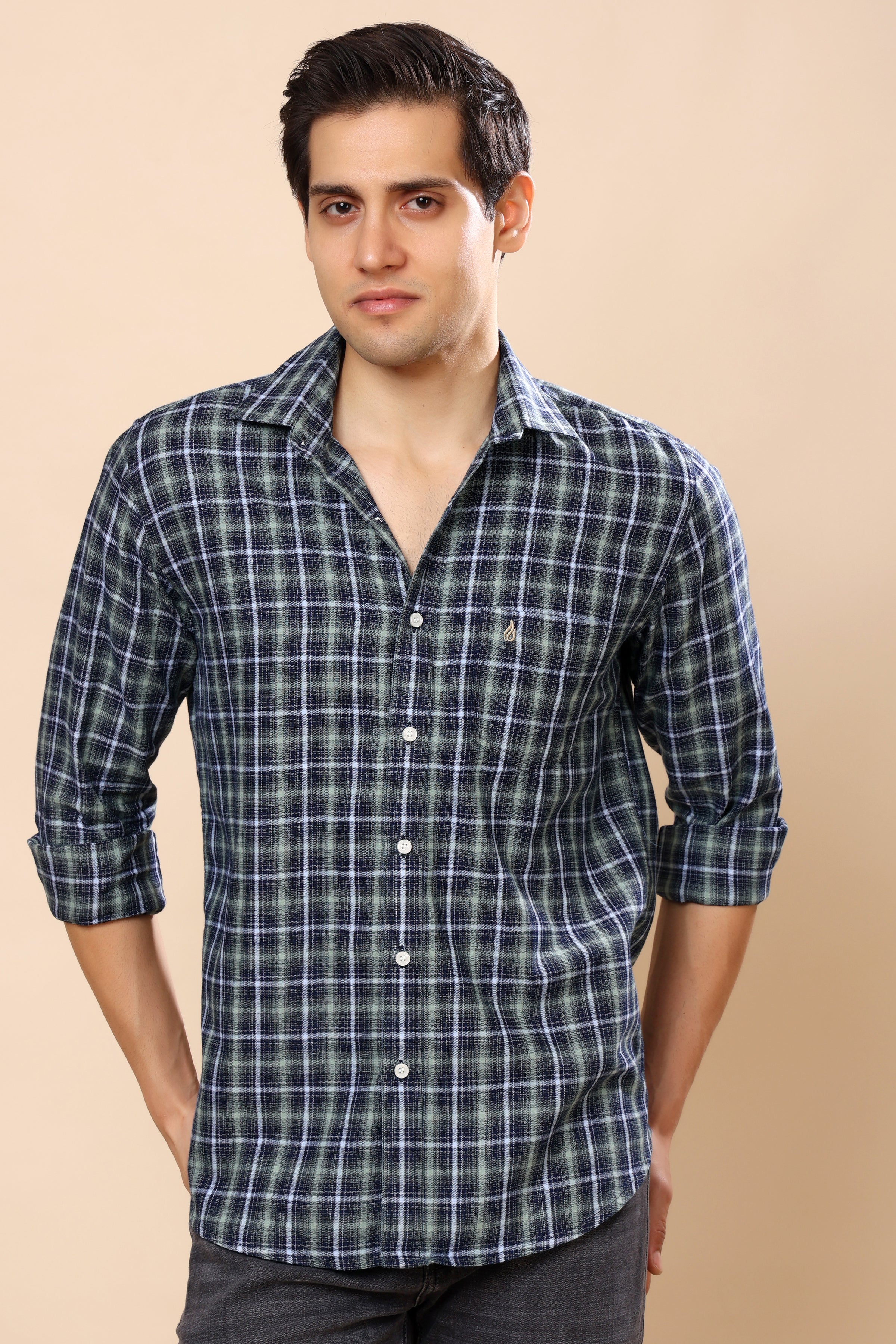 A man wearing a long-sleeve plaid button-up shirt in dark green and white, with the sleeves rolled up, set against a solid beige background.