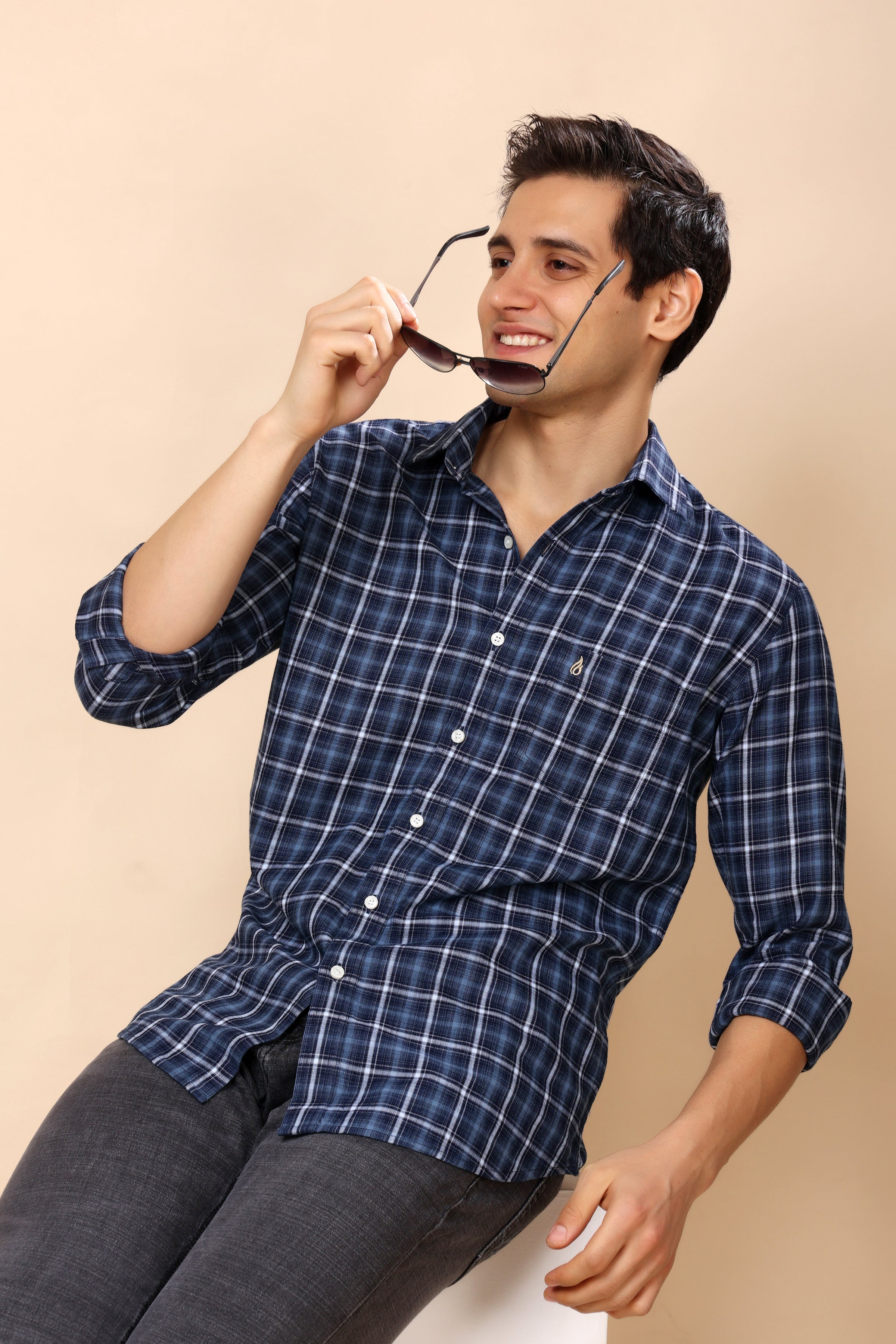 A man wearing a navy blue and white plaid button-down shirt, holding a pair of sunglasses near his face, seated against a solid beige background.