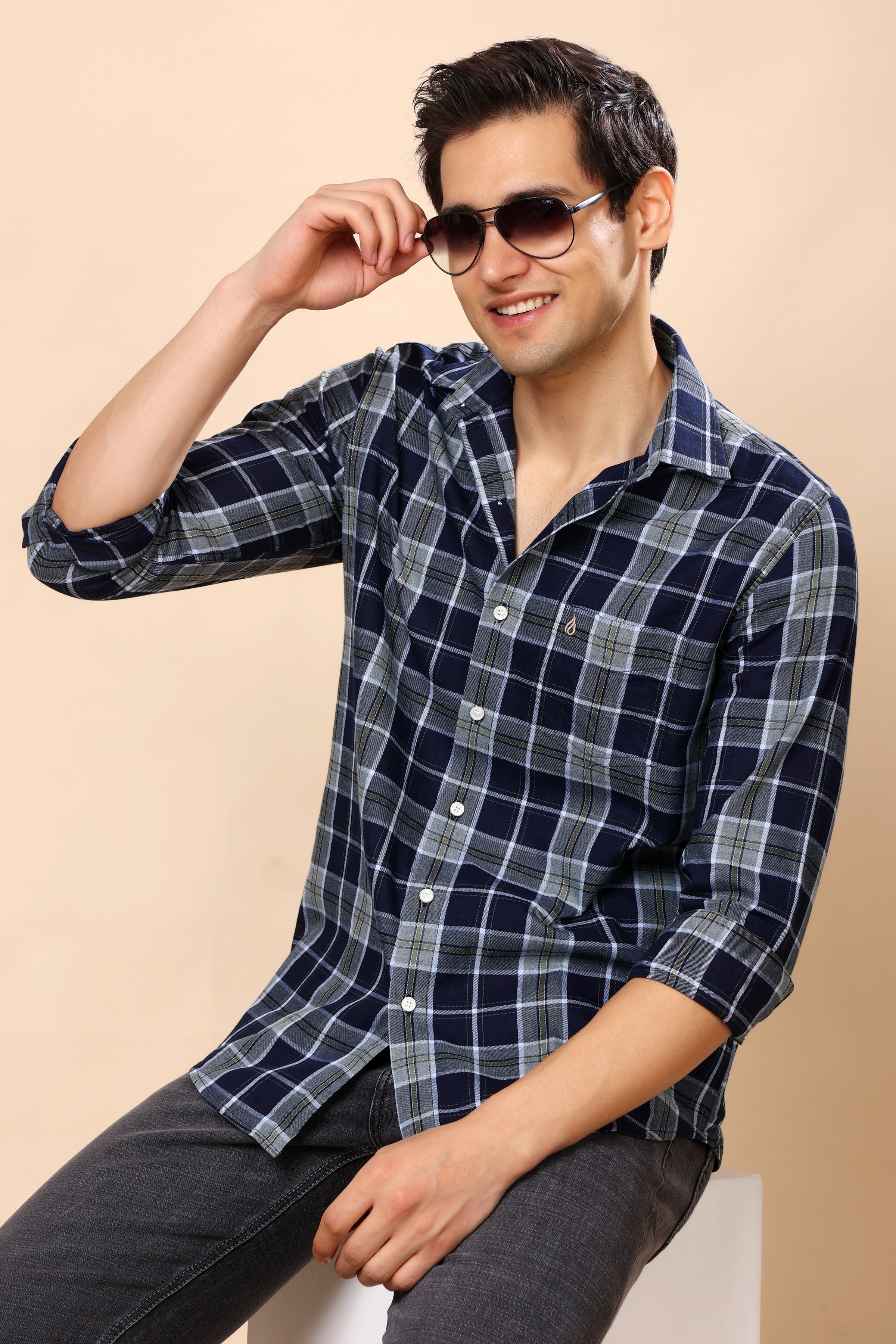 A man wearing a navy blue and gray plaid button-down shirt with dark gray jeans, sitting against a solid beige background. He is wearing dark sunglasses and holding one up to his eye.
