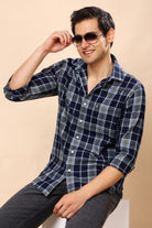 A man wearing a navy blue and gray plaid button-down shirt with dark gray jeans, sitting against a solid beige background. He is wearing dark sunglasses and holding one up to his eye.