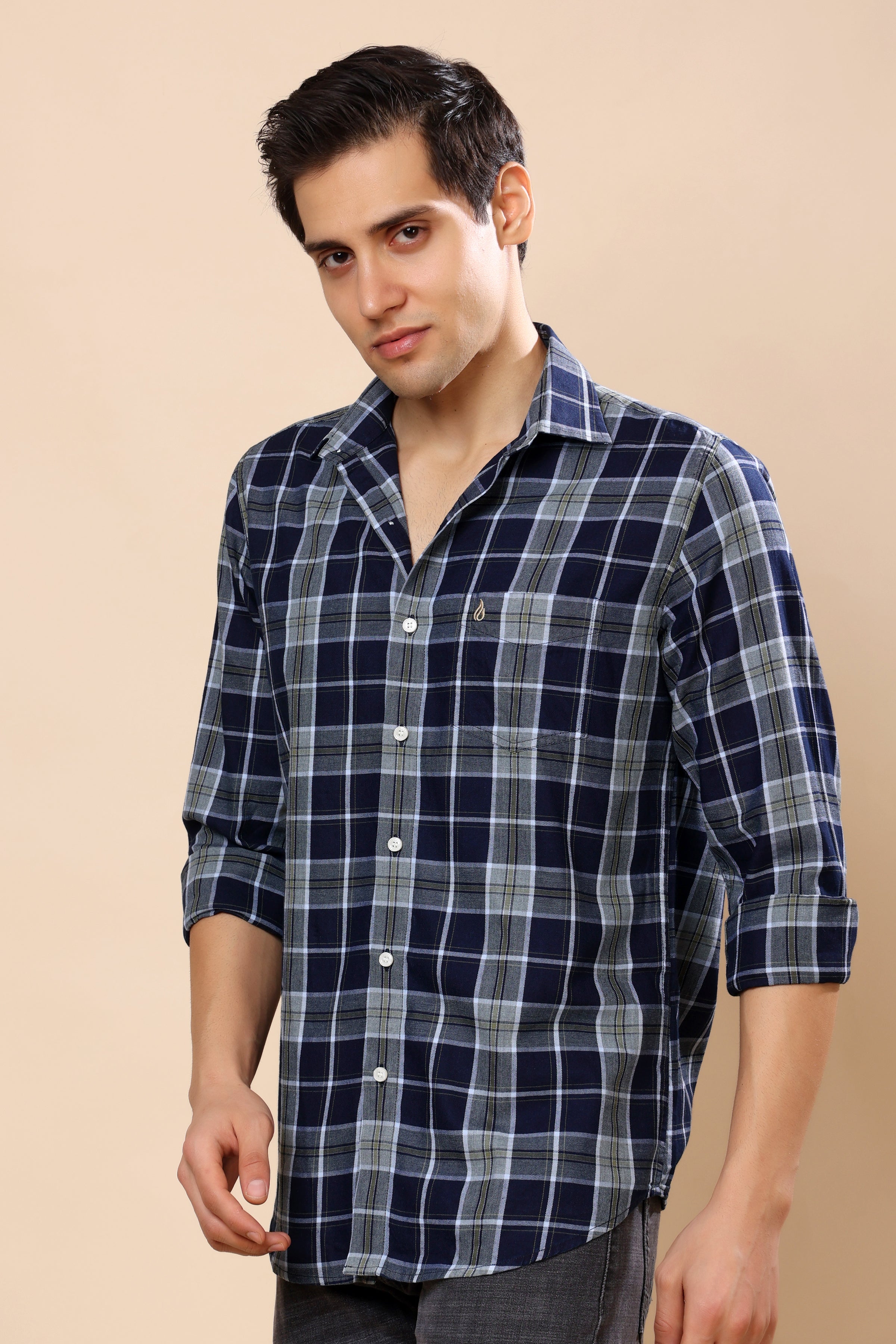 A man wearing a navy blue and gray plaid long-sleeve button-down shirt, standing against a solid beige background.