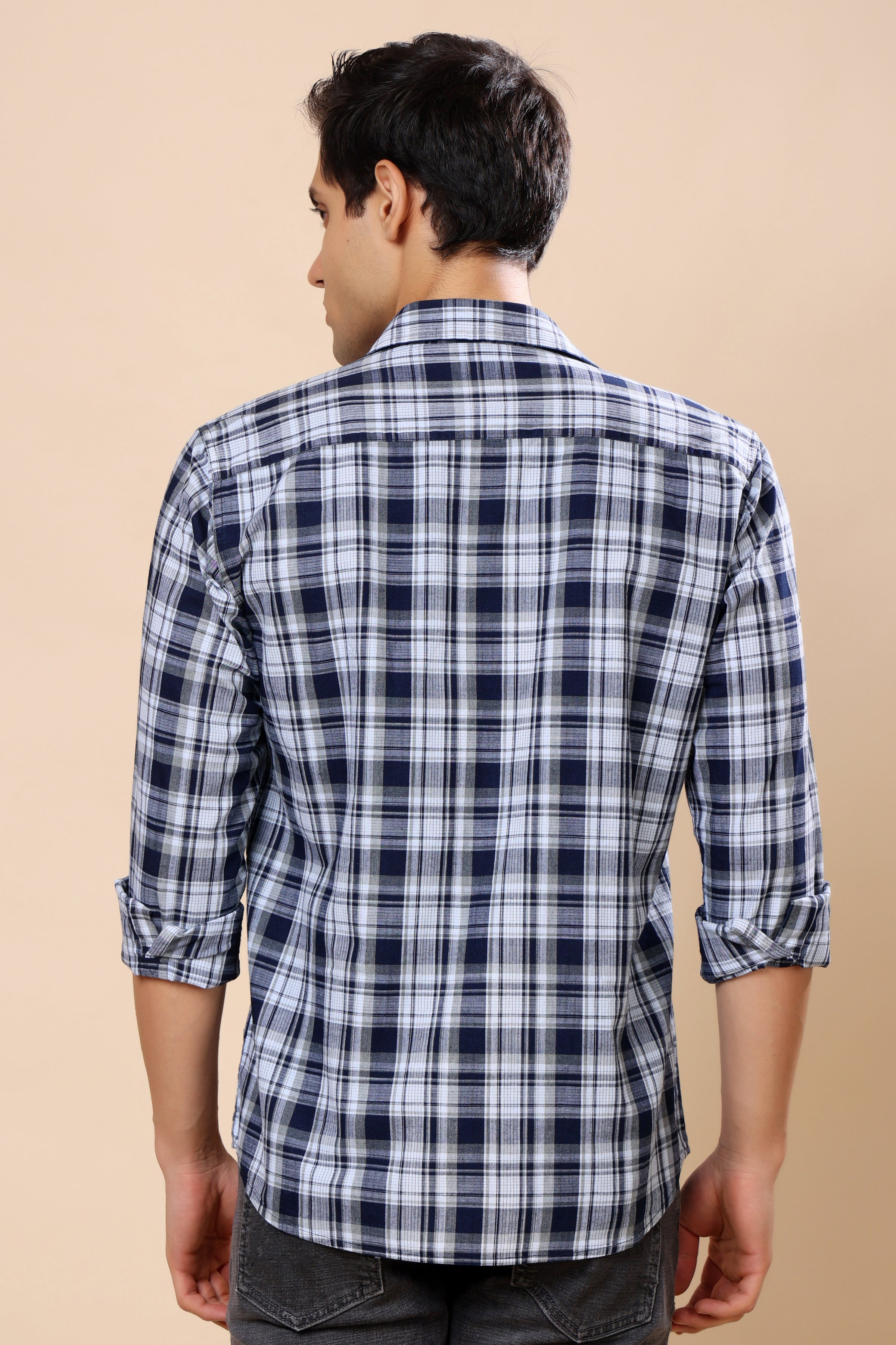 A man viewed from the back wearing a navy blue and white plaid long-sleeve shirt with rolled-up sleeves, set against a solid beige background.