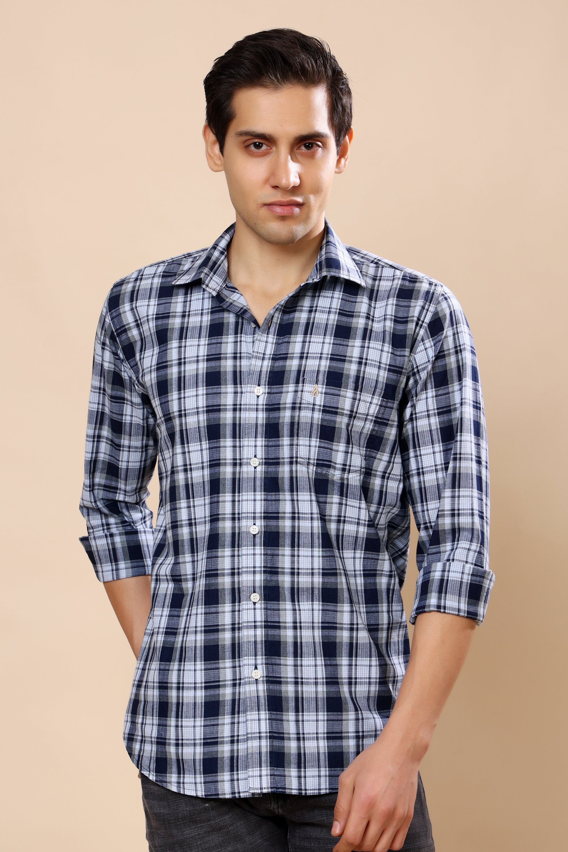 A man wearing a long-sleeve navy blue and white plaid button-down shirt, standing against a solid beige background.