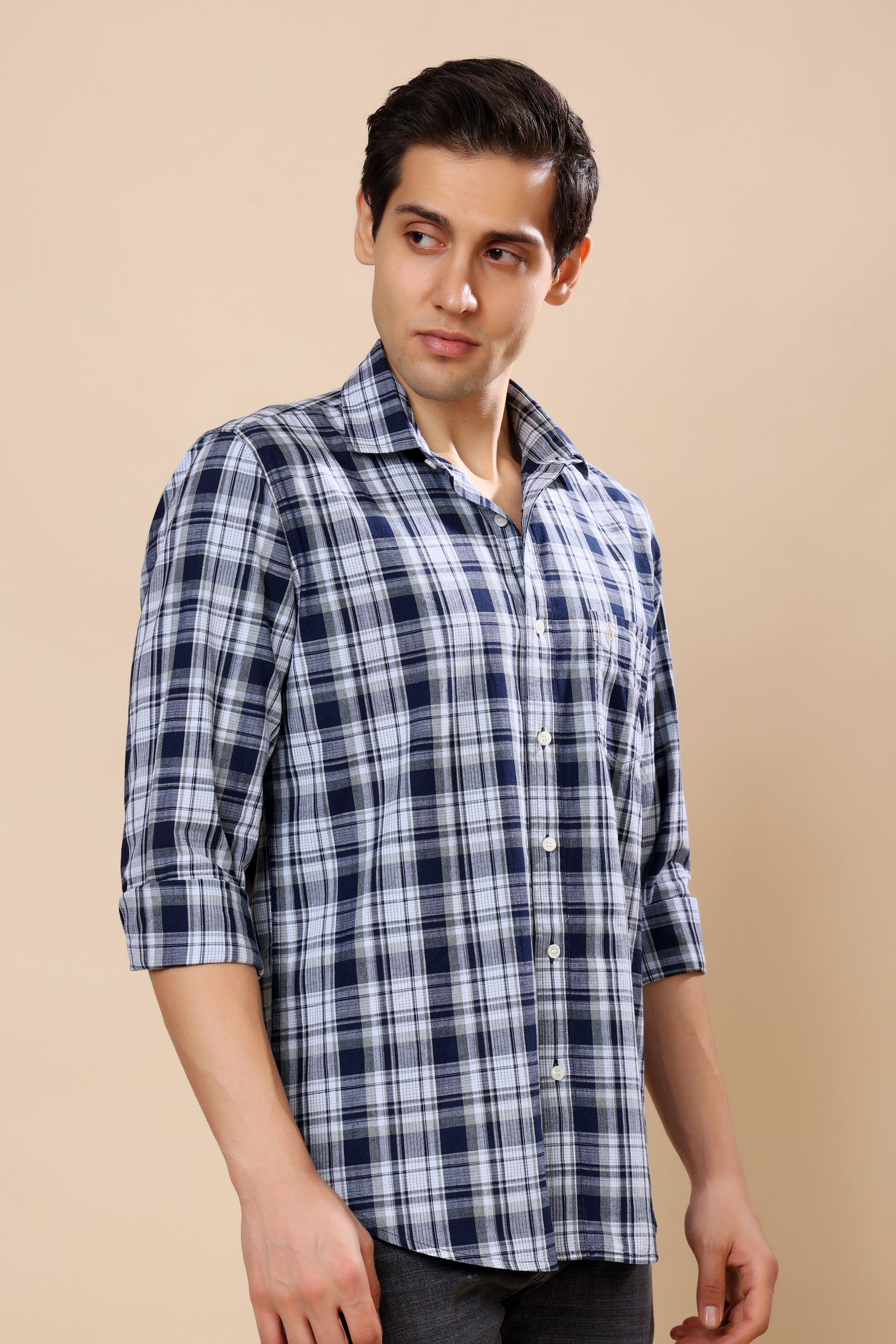 A man wearing a blue and white plaid button-down shirt with rolled-up sleeves, standing against a solid beige background.