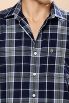 A close-up of a men's plaid button-down shirt worn by a model. The shirt features a navy blue, gray, and white checkered pattern with a small gold embroidered flame logo on the left chest.