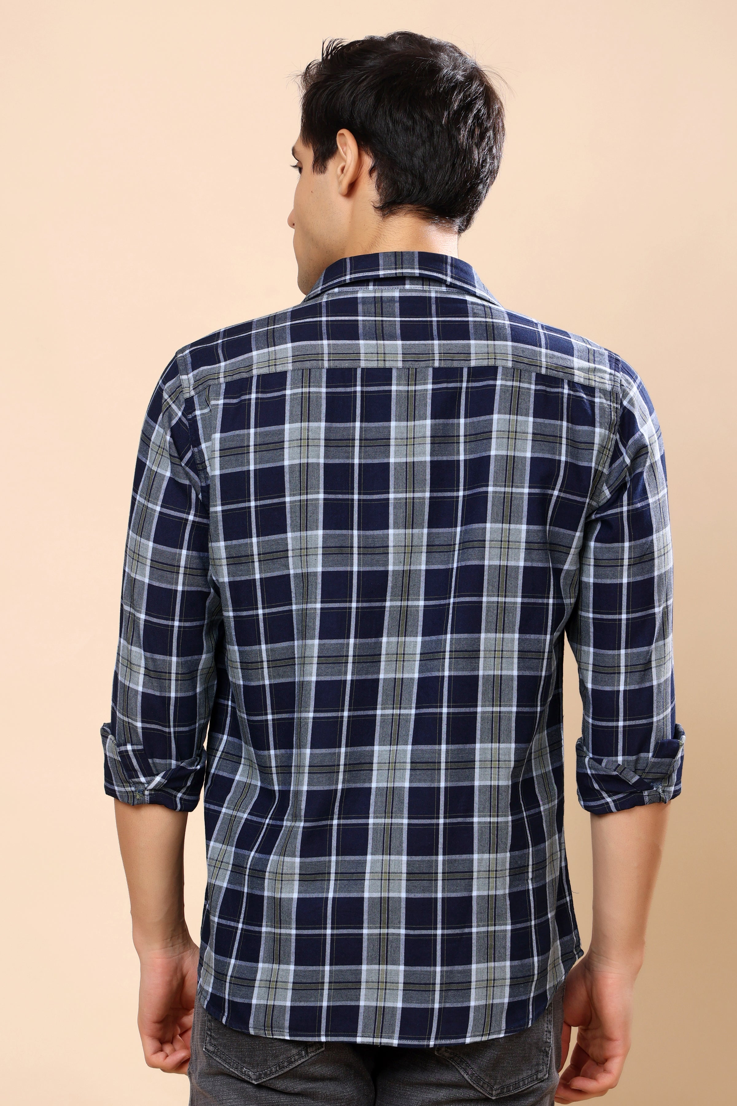 A man viewed from the back wearing a navy blue and gray plaid long-sleeve shirt with rolled-up sleeves, set against a solid beige background.