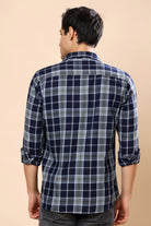 A man viewed from the back wearing a navy blue and gray plaid long-sleeve shirt with rolled-up sleeves, set against a solid beige background.