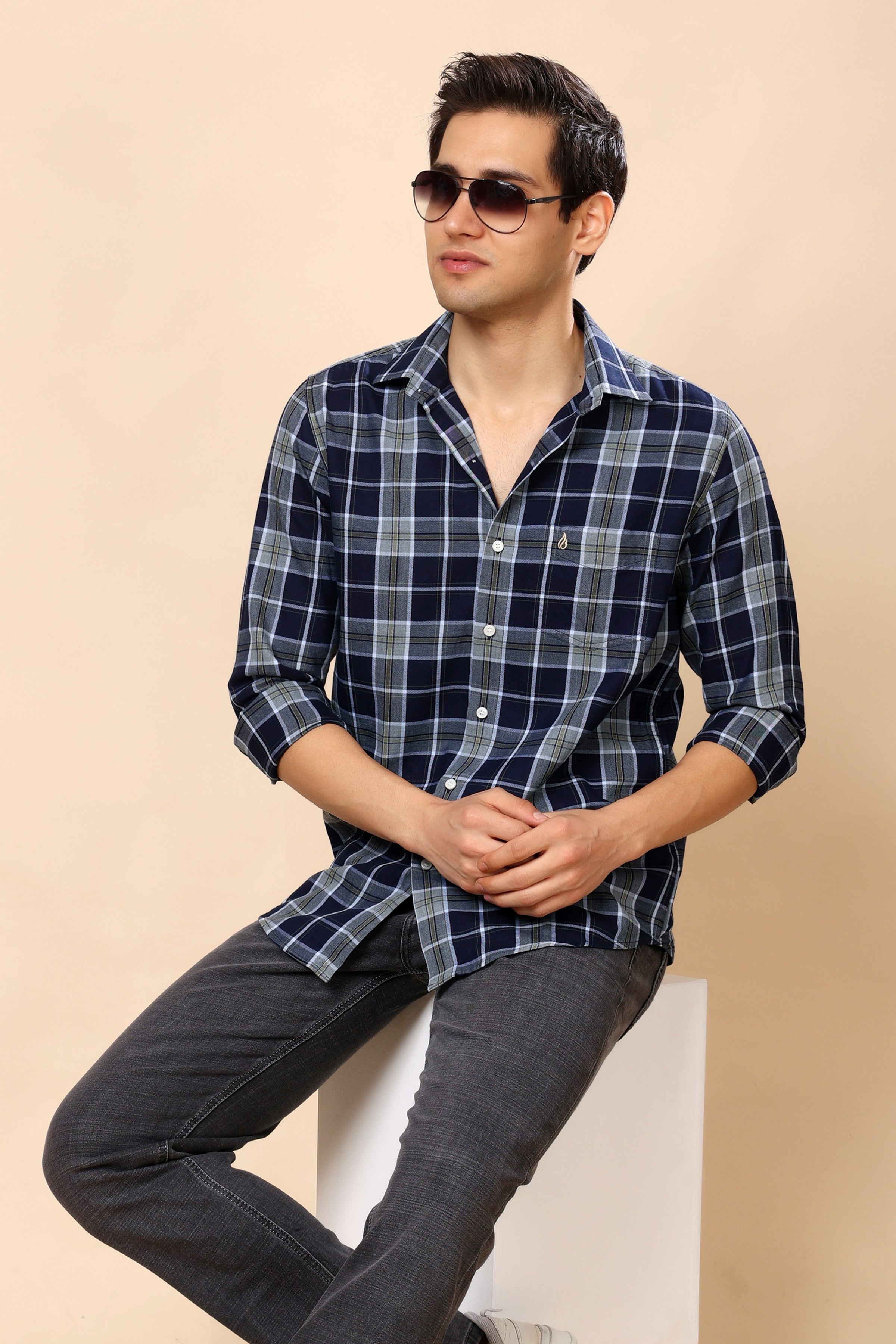 A man wearing a navy blue and gray plaid button-down shirt with dark gray jeans, sitting on a white block against a beige background.