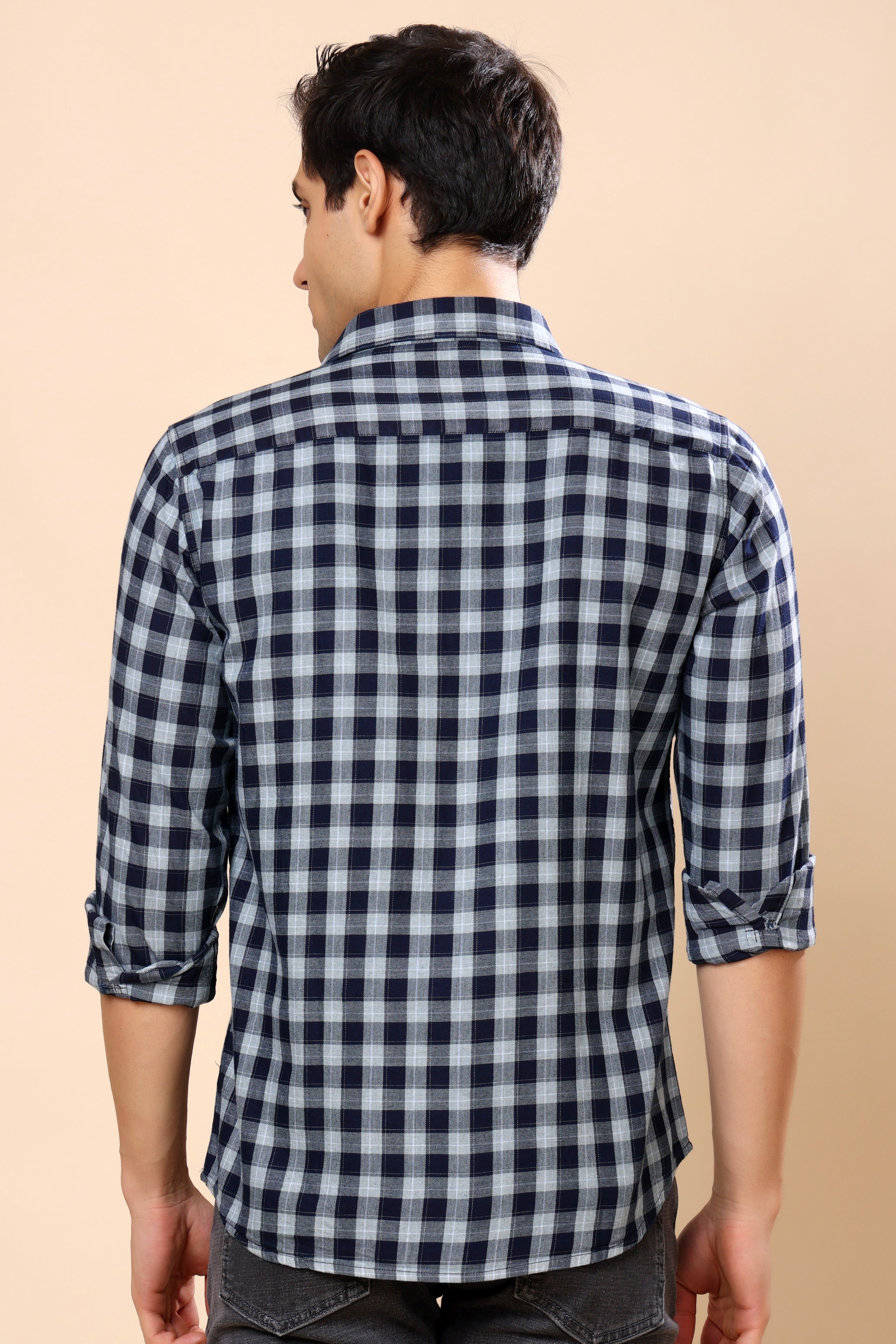 A man viewed from the back wearing a navy and white checkered long-sleeve shirt with rolled-up sleeves, paired with dark gray jeans, against a solid beige background.