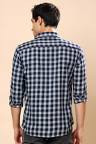 A man viewed from the back wearing a navy and white checkered long-sleeve shirt with rolled-up sleeves, paired with dark gray jeans, against a solid beige background.