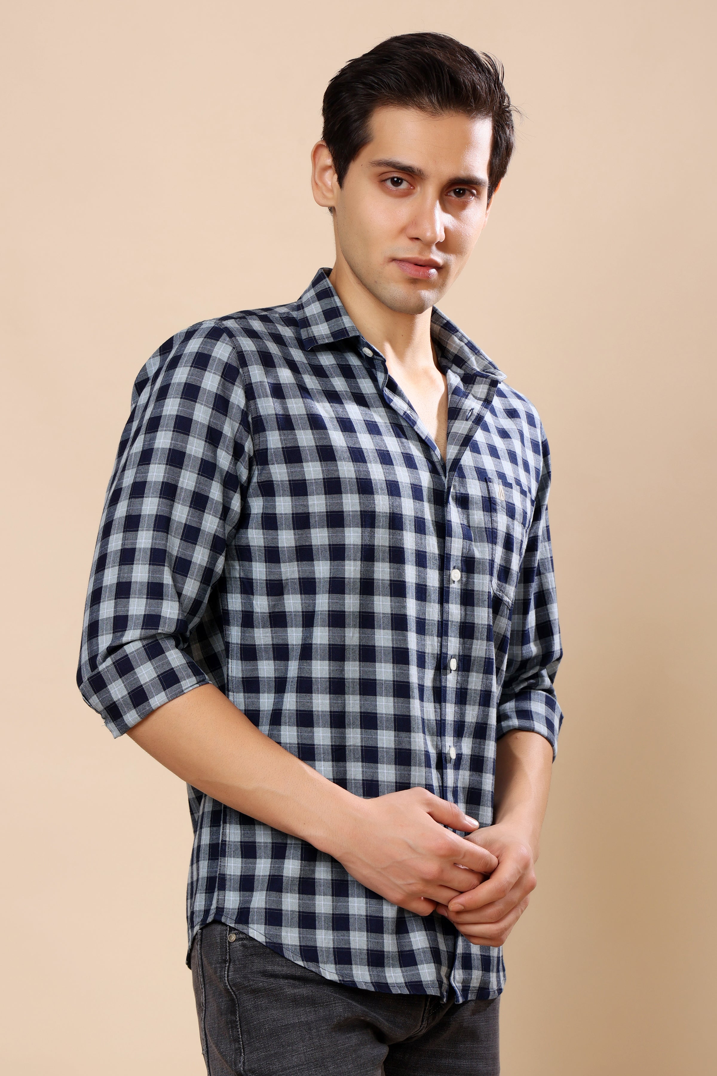 A man wearing a black and white gingham check long-sleeve button-down shirt, paired with dark gray jeans, standing against a solid beige studio background.