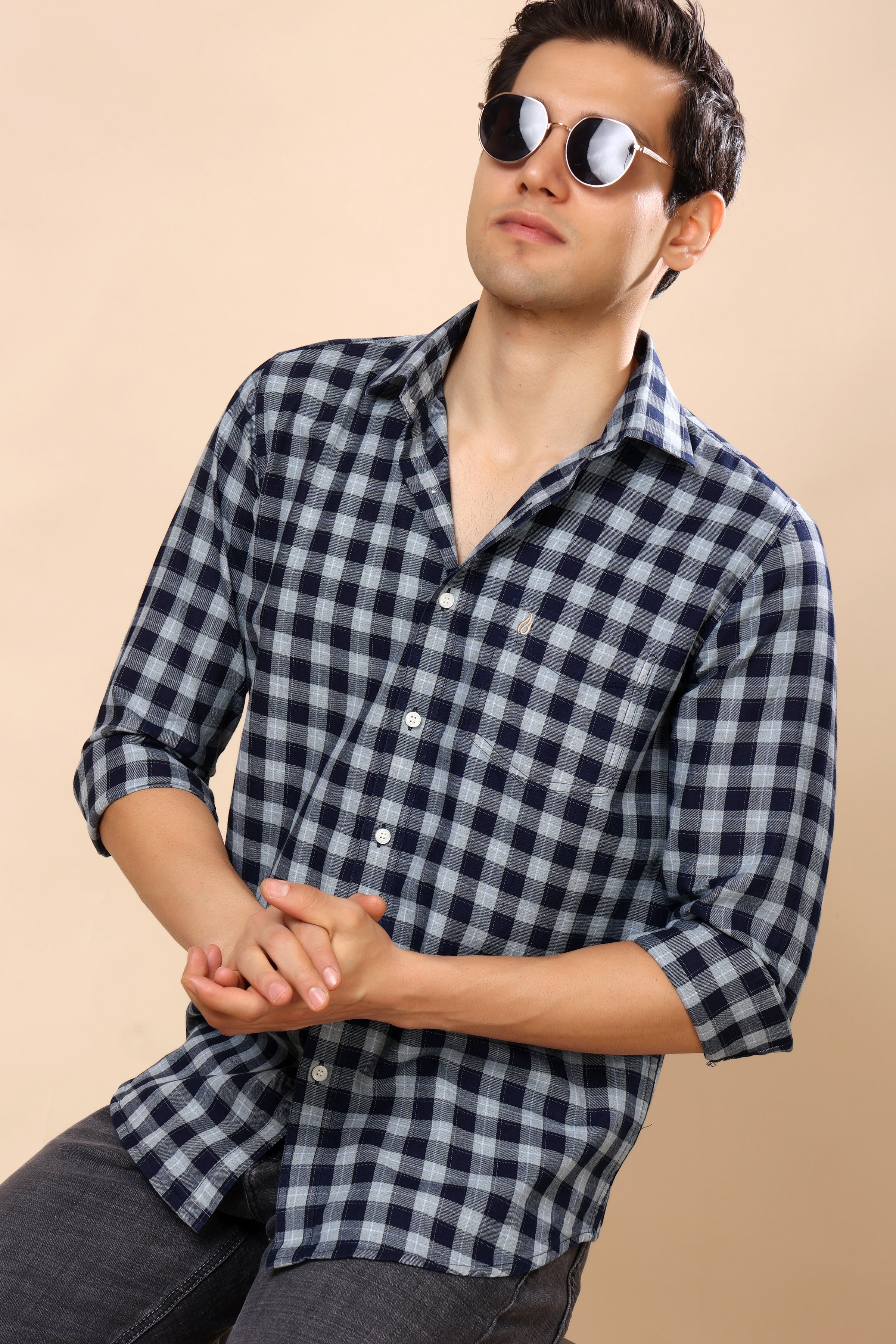 A man wearing a navy and gray checkered long-sleeve button-up shirt, dark gray jeans, and round sunglasses, posed against a solid beige background.