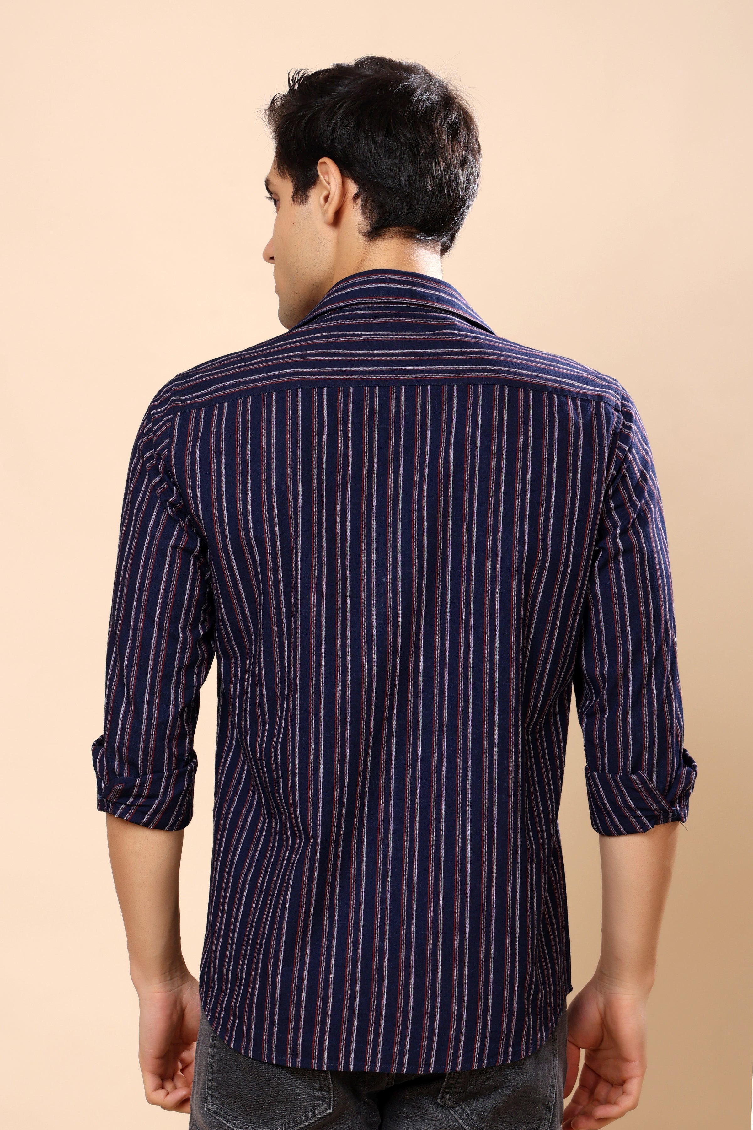 A man viewed from the back wearing a navy blue striped button-down shirt with thin red and white vertical lines, set against a solid beige background.