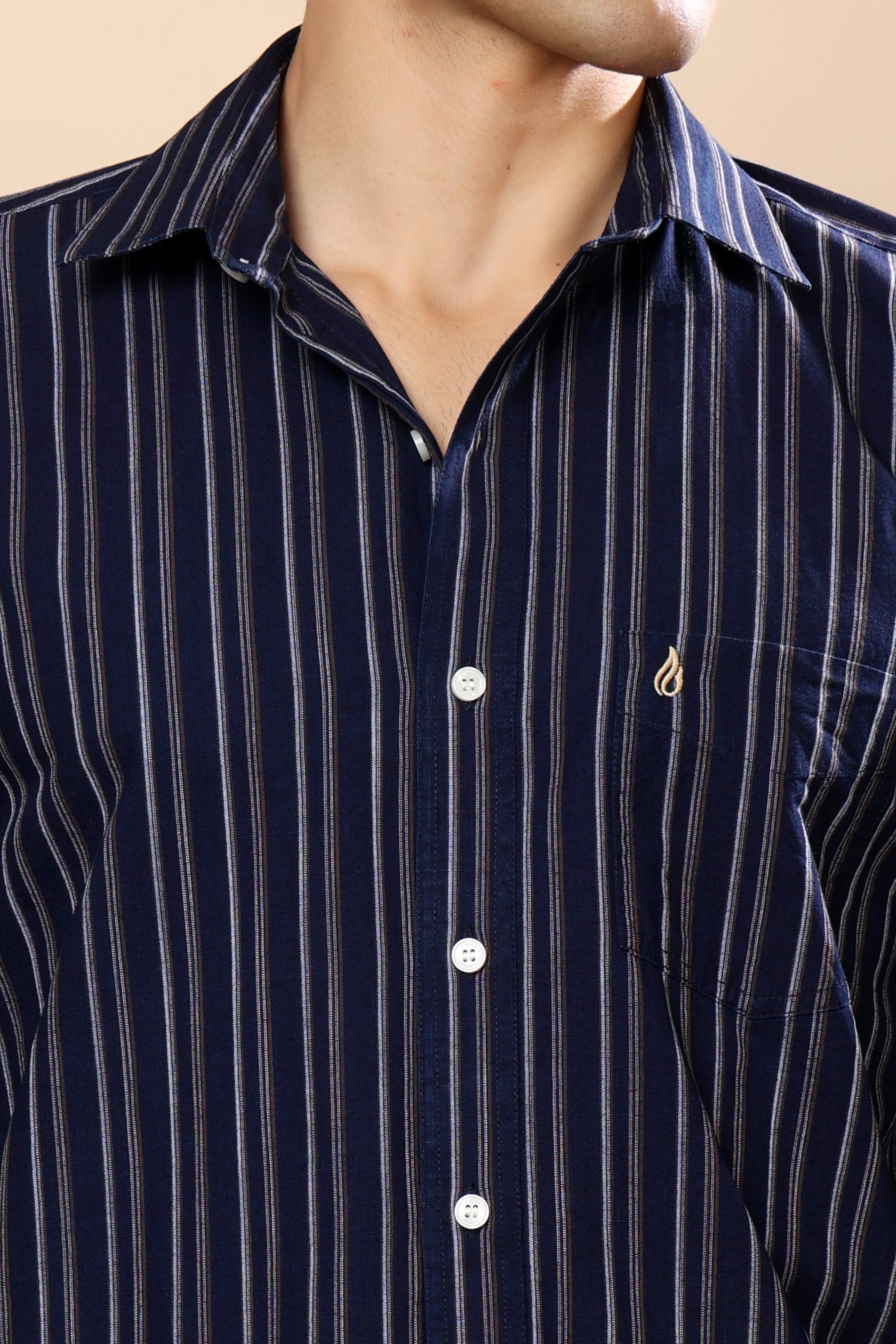 A close-up of a man wearing a navy blue button-down shirt with thin vertical white stripes and a small gold embroidered logo on the chest pocket.