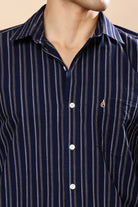 A close-up of a man wearing a navy blue button-down shirt with thin vertical white stripes and a small gold embroidered logo on the chest pocket.