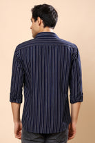 A man viewed from the back wearing a navy blue and white vertically striped long-sleeve shirt, paired with dark gray jeans, against a solid beige background.