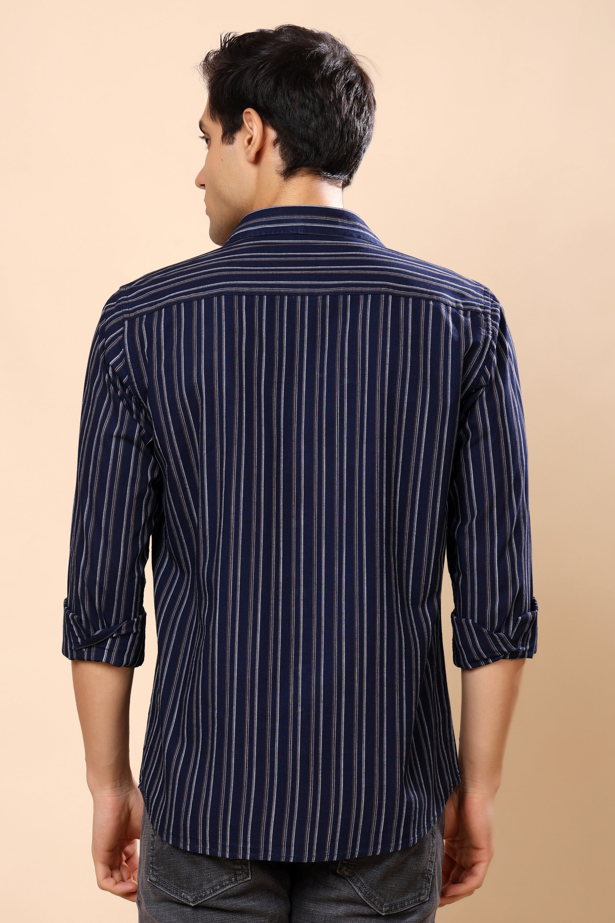 A man viewed from the back wearing a navy blue and white vertically striped long-sleeve shirt, paired with dark gray jeans, against a solid beige background.