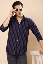 A man wearing a navy blue and purple vertically striped button-down shirt with white buttons, paired with dark gray jeans and white-framed sunglasses, standing against a beige background.