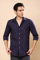 A man wearing a navy blue and purple vertically striped long-sleeve button-down shirt, standing against a solid beige background.