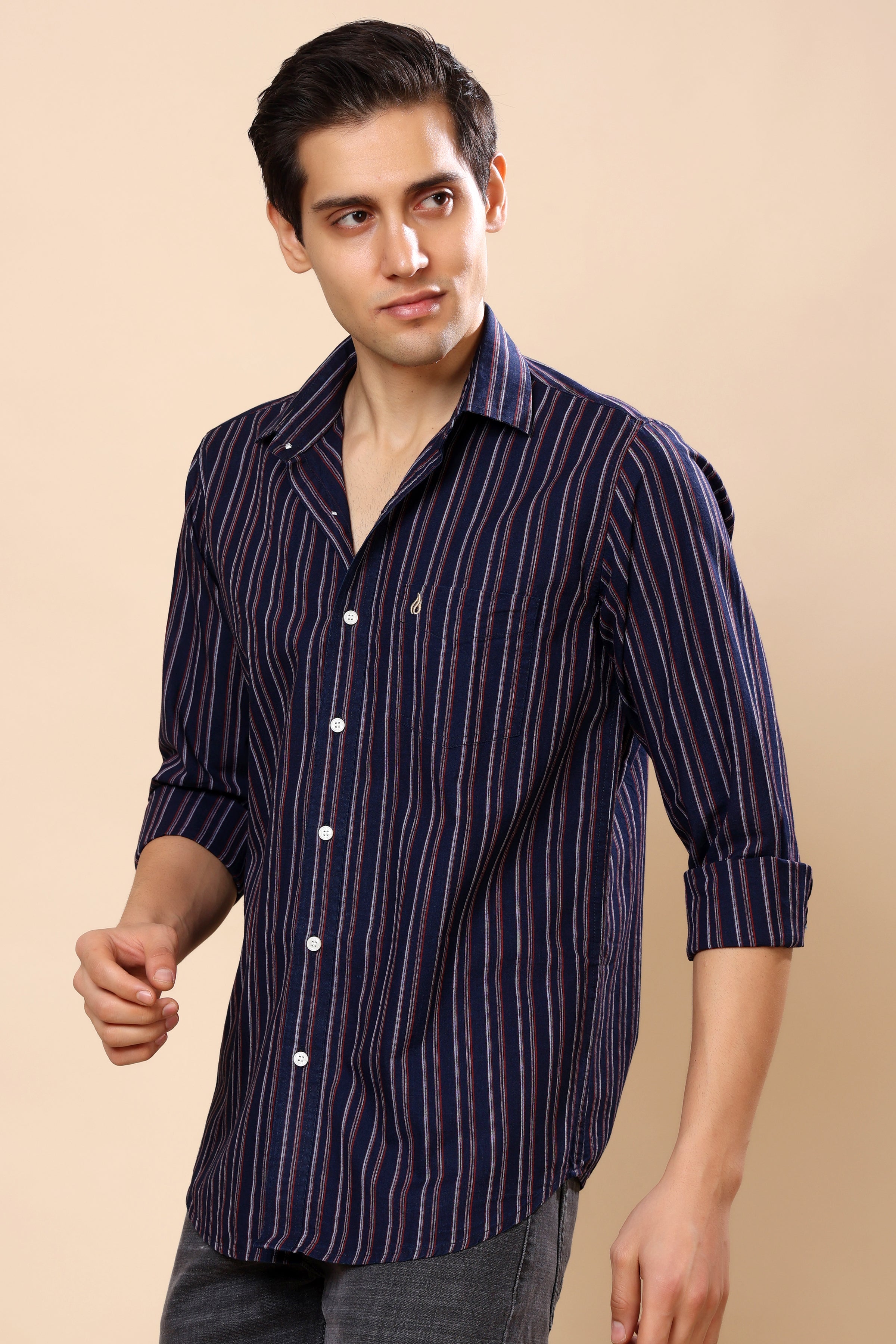 A man wearing a navy blue and black vertically striped long-sleeve button-down shirt, paired with gray jeans, against a solid beige background.