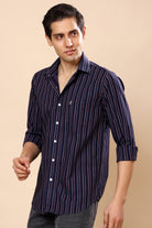 A man wearing a navy blue and black vertically striped long-sleeve button-down shirt, paired with gray jeans, against a solid beige background.