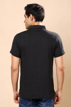 A man viewed from the back wearing a short-sleeve black textured polo shirt and blue jeans, set against a solid beige studio background.