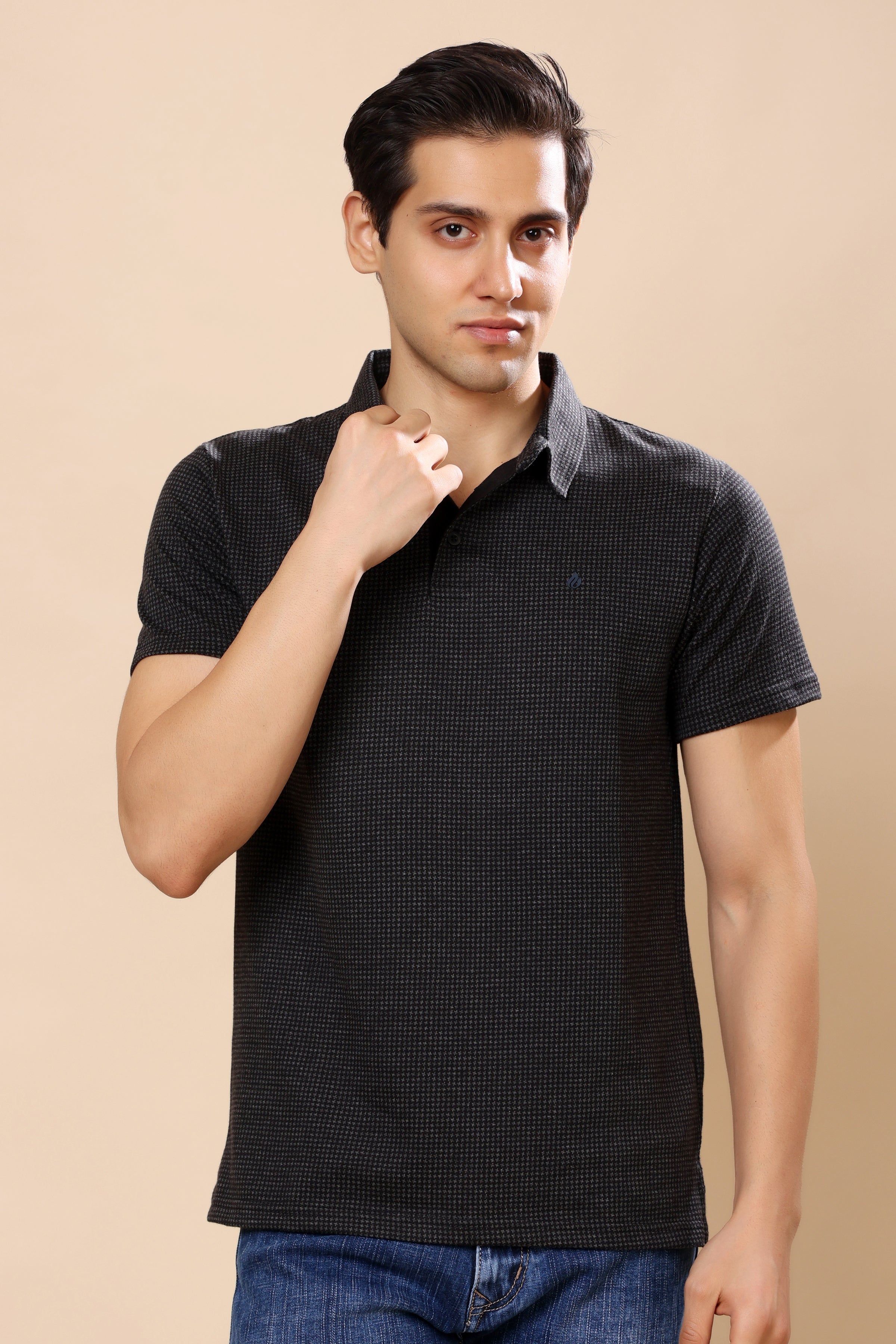 A man wearing a dark charcoal gray textured short-sleeve polo shirt with a contrasting light gray inner collar, paired with blue jeans, set against a solid beige background.