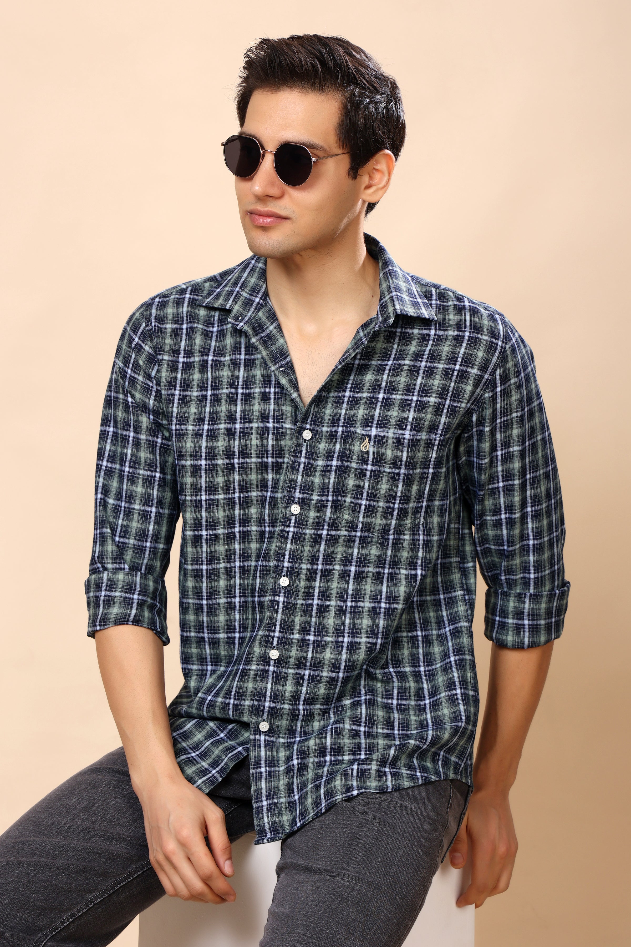 A man wearing a dark blue and green plaid button-down shirt with rolled-up sleeves, paired with dark gray jeans and round sunglasses, seated against a solid beige background.