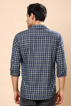 A man viewed from the back wearing a long-sleeve plaid button-up shirt in dark blue and white, paired with dark gray jeans, against a solid beige background.