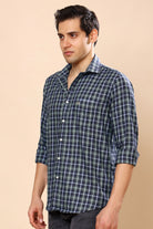 A man wearing a dark green and navy blue plaid button-down shirt with white lines, standing against a solid beige background.