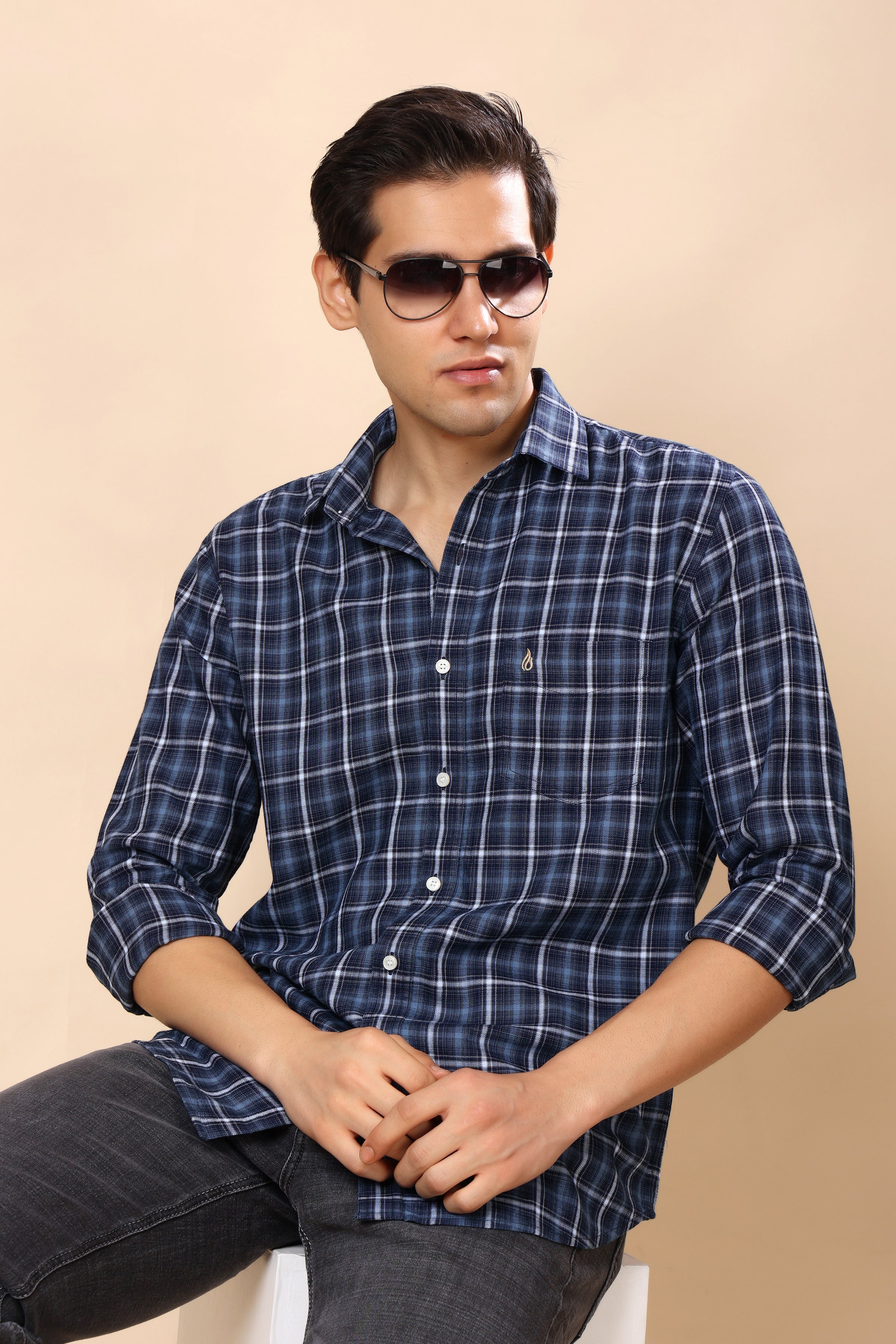 A man wearing a navy blue and white plaid button-down shirt with dark sunglasses, sitting against a solid beige background.